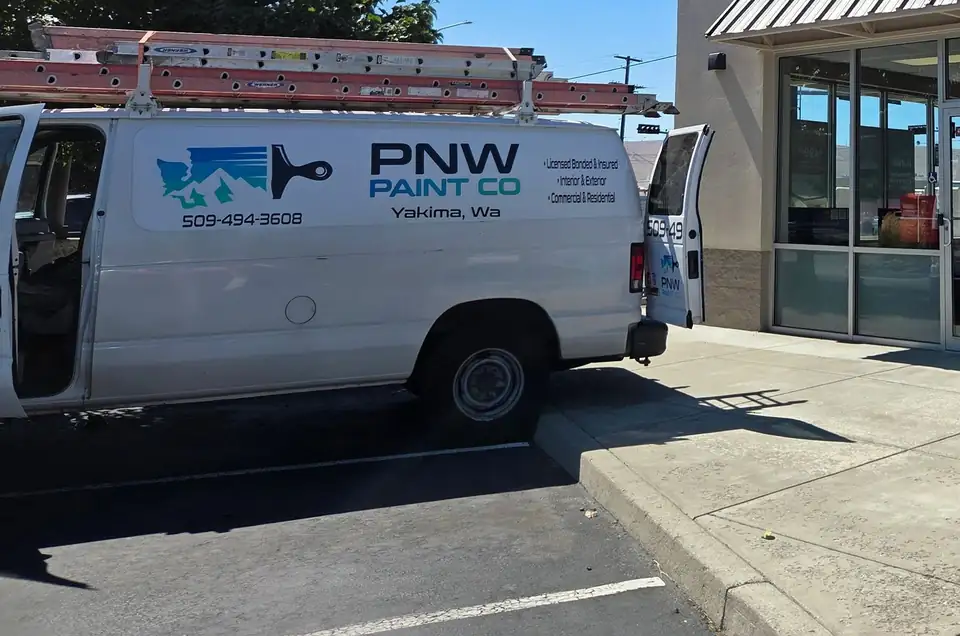 PNW Paint Company work van with the company logo, phone number and ladders on the roof rack, parked at a storefront job